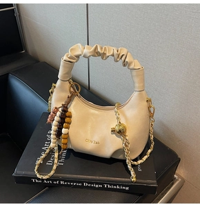 Detail of beige pleated handbag with beaded charm and gold chain on leather surface