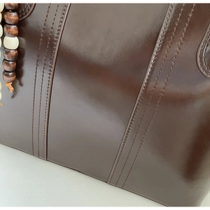 Dettaglio borsa tote in pelle marrone scuro lucido con cuciture precise e ciondolo in legno
