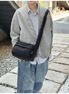 Outdoor street shot of a black nylon crossbody bag with wide strap and orange label detail