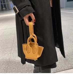 Street style shot of a yellow litchi leather mini bucket bag held by a model in black