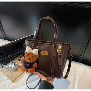 Detail of dark brown tote bag with metal plaque, teddy bear charm and office accessories