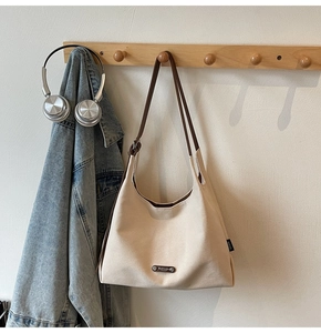 Detail view of beige canvas tote bag with brown straps and metal logo on a wooden hanger