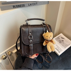 Close-up of a dark brown matte leather crossbody bag featuring dual buckle straps and a bear charm