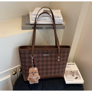 Brown checkered tote bag with plush dog charm and silver logo detail on bookshelf