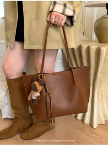 Detail of a brown leather tote bag with a knitted bear charm and long slim handles