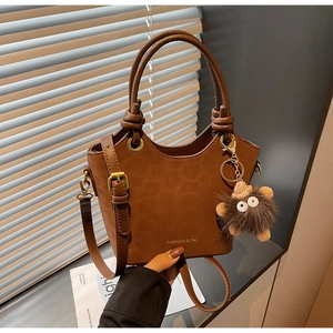 Dettaglio borsa a secchiello marrone opaco con testo oro, finiture ottone e ciondolo peluche