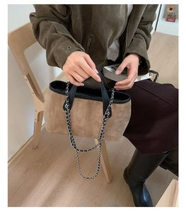 Detail of a brown suede bucket bag featuring black handles and a metallic woven chain strap