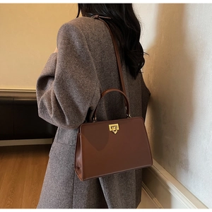 Detailed view of brown leather trapezoid handbag with gold metal lock on a model coat