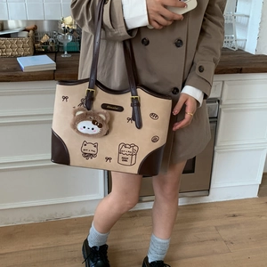 Close-up of beige tote bag with cat illustrations, brown leather trim and plush charm
