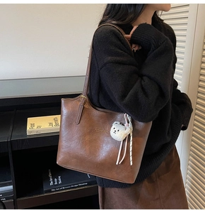 Detail of a brown leather tote bag featuring a brand logo and a white plush bear charm