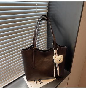 Detail of deep brown tote bag featuring golden brand logo and white plush bear charm