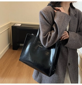 Schwarze Lackleder-Einkaufstasche mit Goldlogo an einem Model mit Tweed-Mantel getragen