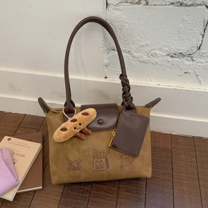 Close-up of khaki tote bag with leather flap and plush baguette pendant on the handle