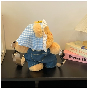 Close-up of plush bear crossbody bag with blue bandana and denim overalls on a desk