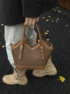 Brown woven tote bag with smooth leather trim and gold buckles in an autumn street setting
