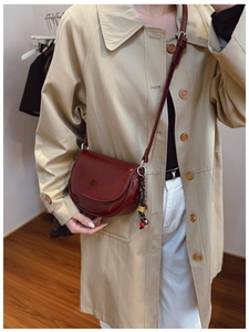 Détail d'un sac besace bordeaux brillant avec breloque porté sur un trench-coat beige