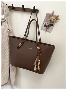 Detail of coffee brown leather tote bag with ethnic bead tassel and gold logo on white wall