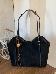 Retro black suede tote bag with adjustable straps and decorative tassel on a wooden desk