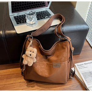 Close-up of brown hobo tote bag with wide strap and plush bear charm on a bench