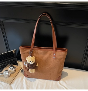 Detail view of brown tote bag with bear embossing, plush charm, and laptop on desk