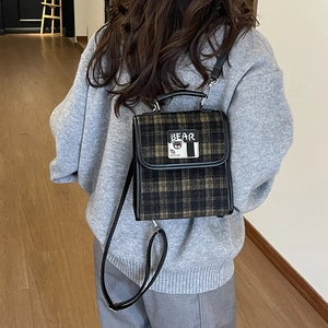Détail du sac à dos à carreaux verts avec écusson ours et lanières sur un pull gris