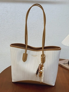 White woven tote bag with brown leather handles and wooden bead tassel on a wooden table