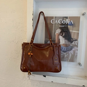 Brown leather tote bag featuring a gold logo and orange beaded tassel charm in a white niche