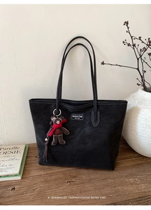 Detail of black suede tote bag with contrast stitching and bear charm on wooden stand