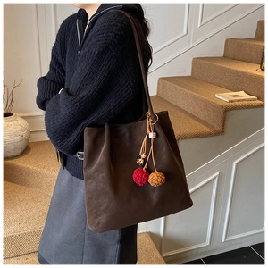 Brown suede tote bag with colorful pompom charm shown on shoulder in interior setting