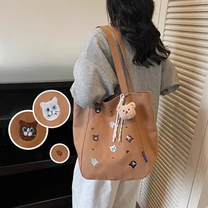Close-up detail of cat head embroidery and plush bear charm on brown canvas tote bag