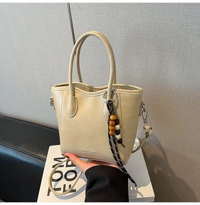 Gros plan du sac seau crème avec un pendentif décoratif en perles et logo frontal