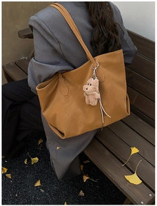Brown leather tote bag with a cute plush bear charm, shown in an autumn outdoor setting