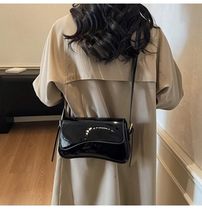 Minimalist black patent leather shoulder bag with gold hardware worn over a beige coat