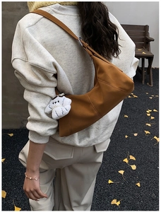 Brown leather crescent bag with white puppy plush charm worn by model in outdoor setting
