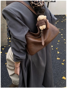 Détail d'un sac hobo en cuir marron avec un charme ours en peluche sur un manteau gris