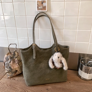 Olive green suede tote bag with golden buckle and a cute plush bunny charm on a wooden table