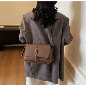 Macro detail of a brown leather shoulder bag with a gold buckle worn with a gray blazer