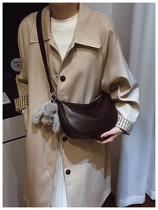 Dark brown crescent leather crossbody bag with gray plush bunny charm on a beige trench coat