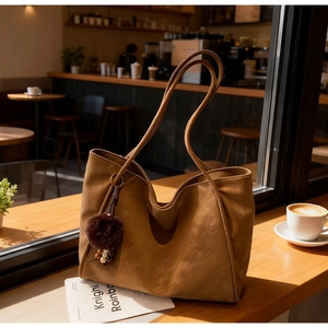 Detail view of a large brown tote bag with a soft plush ball charm in a cozy cafe setting