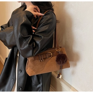 Brown suede shoulder bag with leather buckle detail and fuzzy pompom charm worn with leather coat
