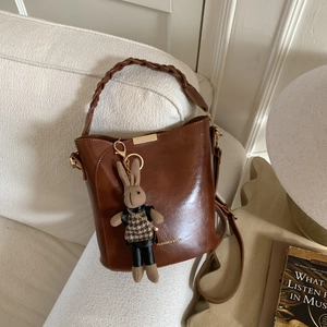 Brown faux leather bucket bag with woven handle and cute rabbit charm on white sofa