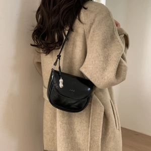 Detail showing a black glossy saddle bag with a silver peanut pendant on a beige coat