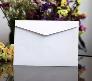 Close-up of minimalist white V-flap envelope standing on mirror surface with floral decor