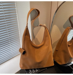 Close-up of caramel leather tote bag with wide strap and cylindrical charm under striped light