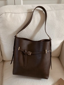 Minimalist dark brown bucket bag in pebbled PU leather with gold buckle detail on sofa