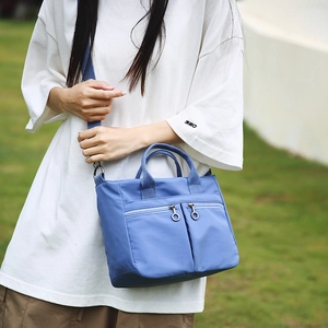 Mist blue nylon tote bag with functional zipper pockets shown in an outdoor casual setting