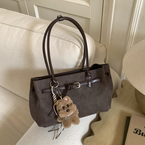 Detail of dark brown suede tote bag with metal buckle and plush bear pendant on sofa