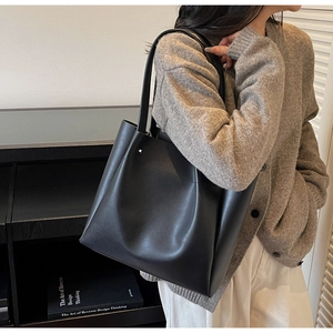 Close-up of a large black minimalist tote bag worn by a model in a casual indoor setting