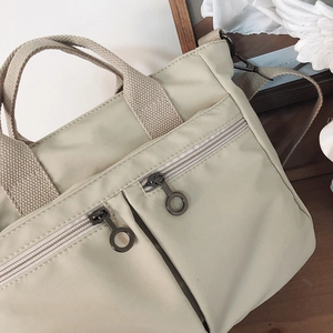 Detail of beige nylon tote bag featuring dual zipper front pockets and metal ring pulls