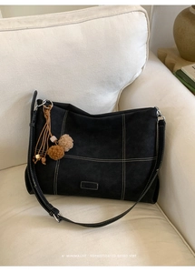 Detail of black suede tote bag with white contrast stitching and plush ball charm on a sofa